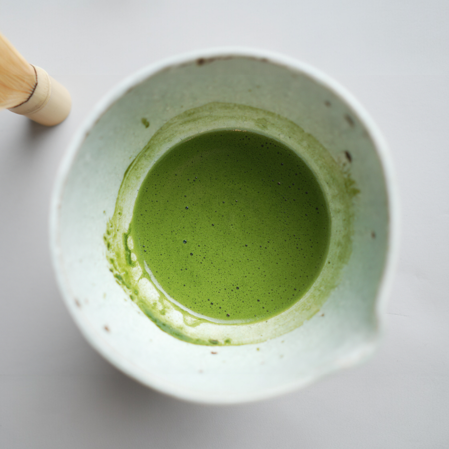 White bowl with green matcha tea and a wooden whisk on a light gray background