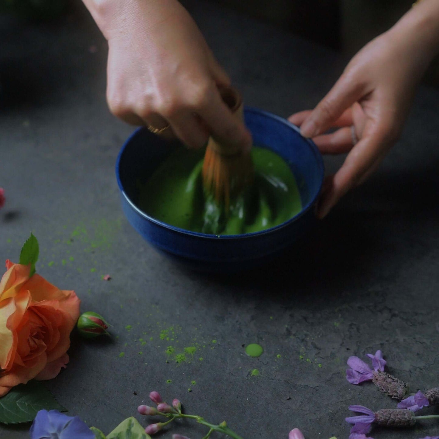 Preparing ceremonial matcha in a blue bowl with a bamboo whisk and fresh flowers surrounding it