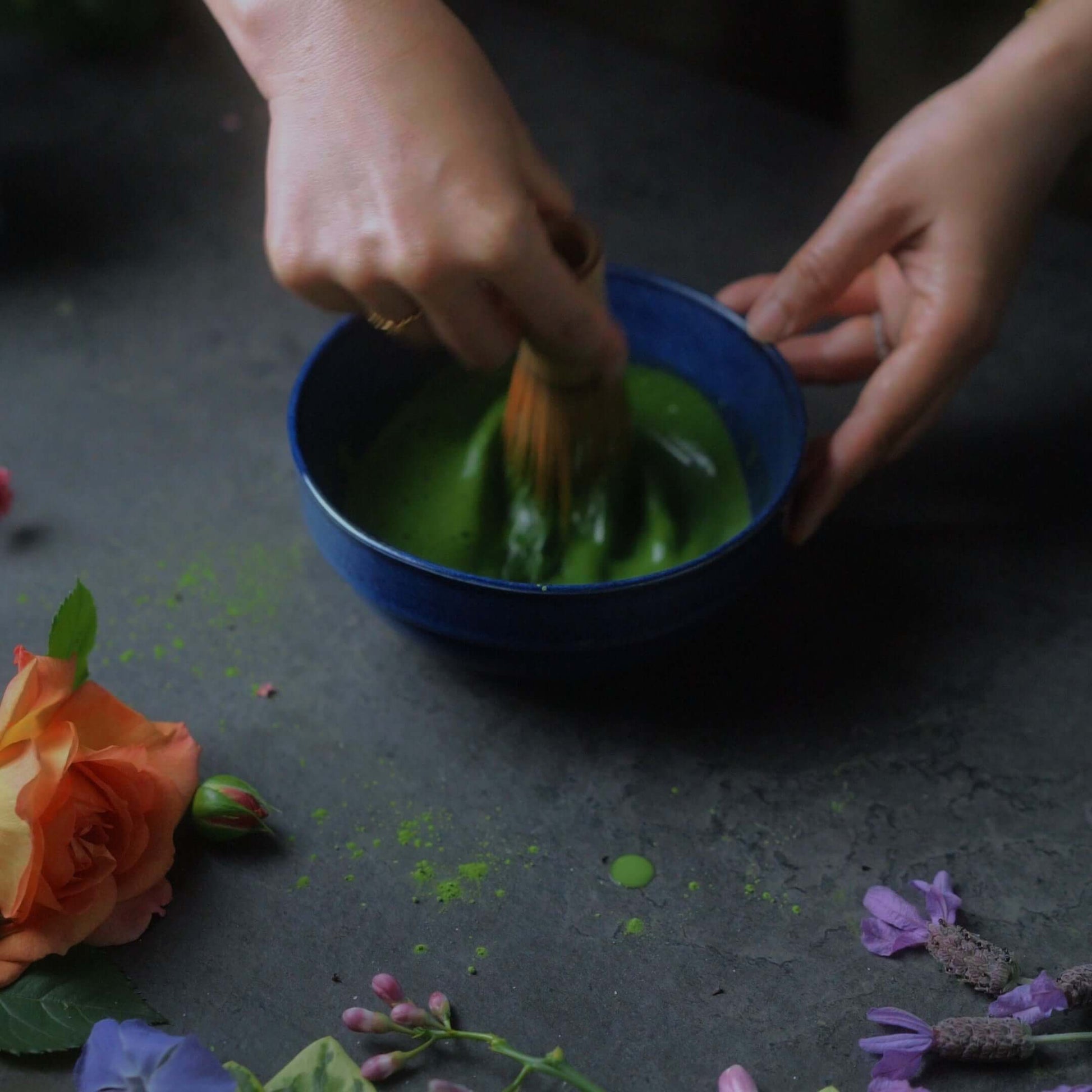 Preparing ceremonial matcha in a blue bowl with a bamboo whisk and fresh flowers surrounding it